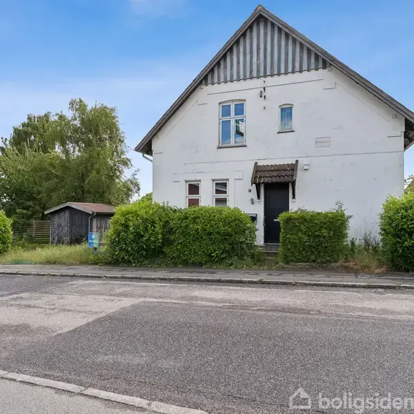 Et hvidt hus med spidst tag står ved en vej. Huset har en lille veranda og omgives af grønne buske. En mindre bygning ses i baghaven.