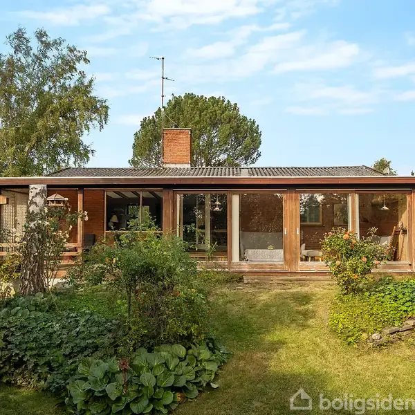 Et enfamiliehus med store vinduespartier står i en velplejet have med græsplæne og forskellige buske. Huset er omgivet af grønne træer og har en skorsten på taget.