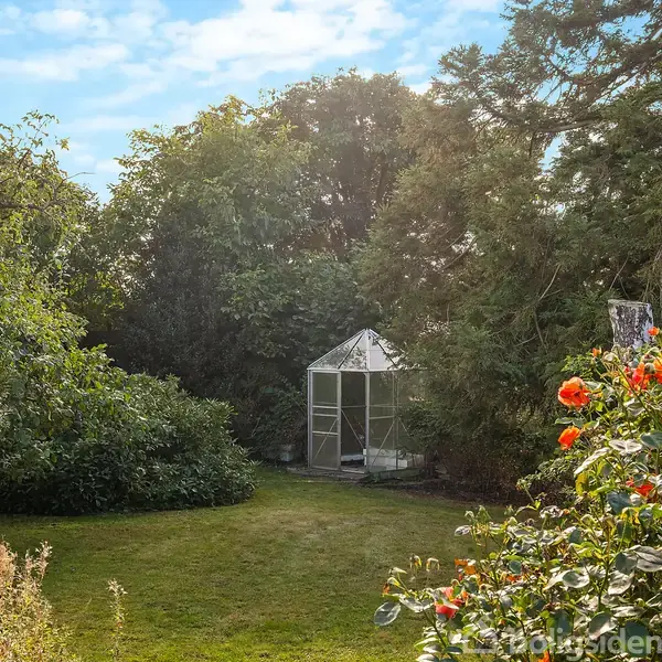 Et lille drivhus står stille omgivet af tætte træer og buske i en frodig have, mens et blomsterbed med røde blomster vokser i forgrunden.