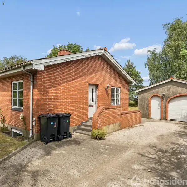 Rødt murstenshus med hvide vinduer står på en brostensbelagt indkørsel. Til højre ses en fritstående garage. Omkring huset er der træer og buske, hvilket skaber en rolig atmosfære.