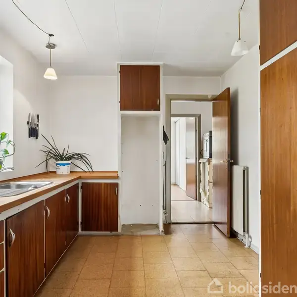 Køkken med mørke træskabe og beige fliser, hvor en vask er placeret under et vindue. To planter står på bordet, og døren fører til en tilstødende gang.