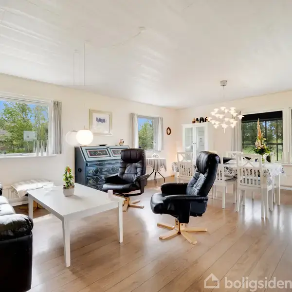 Stue med sorte lænestole og sofa, placeret omkring et hvidt sofabord. I baggrunden ses et spisebord og store vinduer med udsigt til en grøn have.