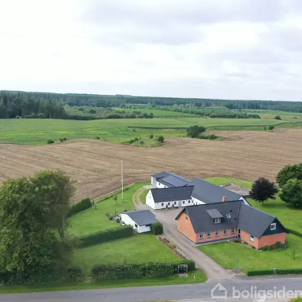 En gård med lange udbygninger ligger omgivet af marker og træer. Bygningerne er røde med grå tage, og en vej fører op til gården fra forgrunden.