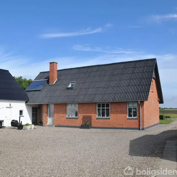 En rødstensbygning med sort tag står på en gårdsplads med grus. Ved siden af er der en hvid bygning. Landskabet i baggrunden har åbne marker under en blå himmel.