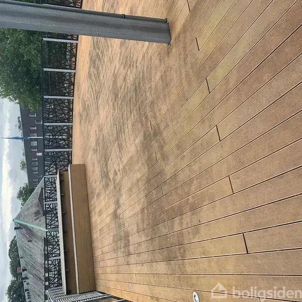 En stor træterrasse strækker sig mod horisonten med en pergola i baggrunden og et grønt område ved siden af. Himlen er overskyet, skaber en stille atmosfære.