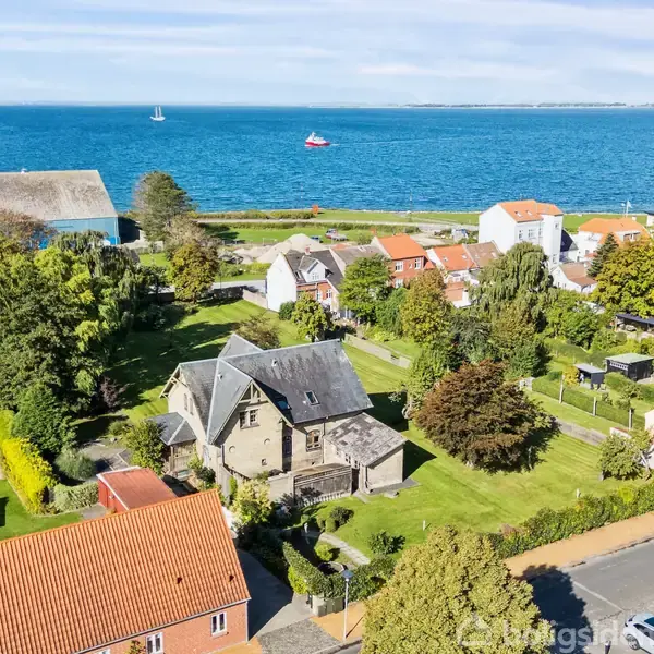 Et hus omgivet af frodig have beliggende nær kystlinjen. Havet er roligt med få både i baggrunden. Omkringliggende bygninger med røde tage skaber en idyllisk kystlandsby.