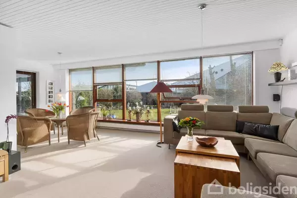 En lys stue med grå hjørnesofa og træbord. Store vinduer lader naturligt lys ind, afslørende en have udenfor. Spisebord med kurvestole i hjørnet.