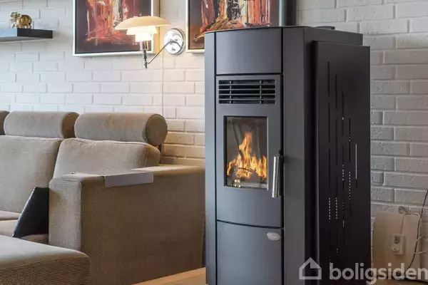 En moderne brændeovn brænder i en hyggelig stue med en grå sofa og hvidmalede murstensvægge. Malerier og en væglampe hænger over sofaen.