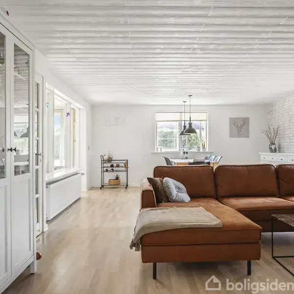 Brun lædersofa står i en lys stue med trægulv, ved siden af en hvid skænk og glasvitrine. Store vinduer og et spisebord i baggrunden giver et rummeligt miljø.