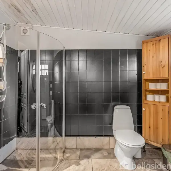 Brusekabine med glasdør, toilet ved siden af og håndvask til venstre. Badeværelset har en sort flisevæg, træloft og træskabe, der indeholder håndklæder. Et vindue lader lys ind.