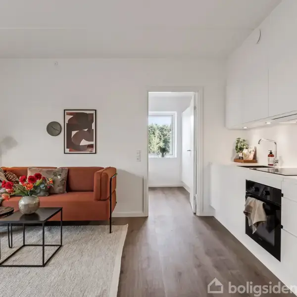 En stue med en orange sofa fyldt med puder og tæpper, ved siden af et lille bord med blomster. Til højre er et hvidt køkken; en dør fører til et andet rum.