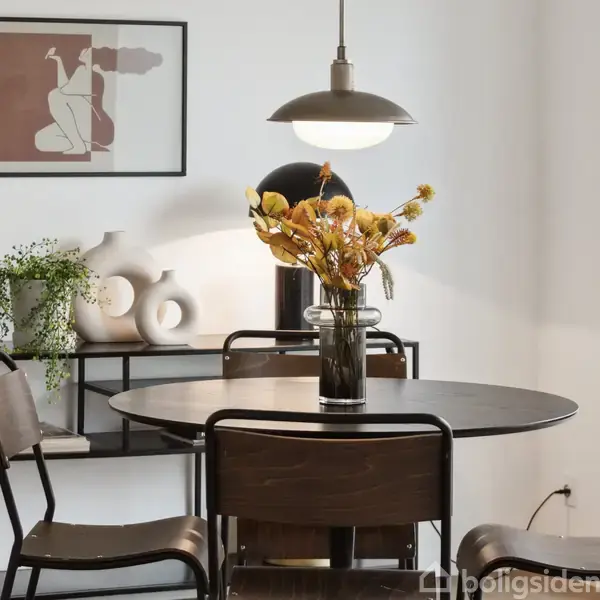 Et rundt bord med fire stole har en vase med tørrede blomster, der står under en hængelampe. I baggrunden er en reol med dekorative vaser og en plante.