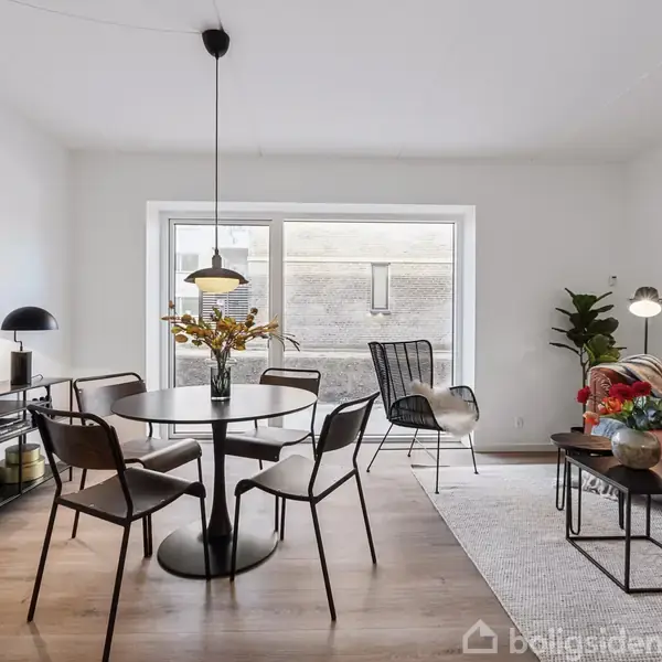 Rundt spisebord med stole står centralt, omgivet af moderne, minimalistiske møbler og lamper. En stor sofa med puder, planter og kunst pryder rummet, som har en lys, åben atmosfære.