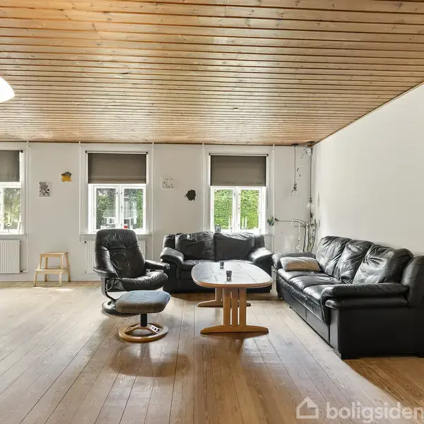 Sort læderhjørnesofa står i en stue med trægulve og træloft. Tre vinduer med gardiner tillader naturligt lys. En stående lampe til venstre oplyser rummet.