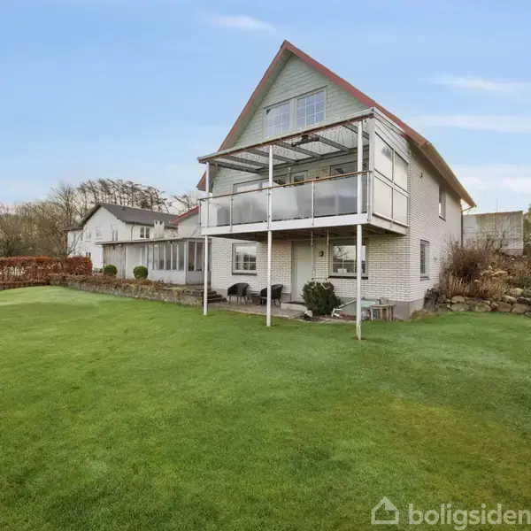 En hvid murstensvilla med to etager og en stor altan. Den står i en velplejet have, omgivet af hække, med en klar blå himmel i baggrunden.