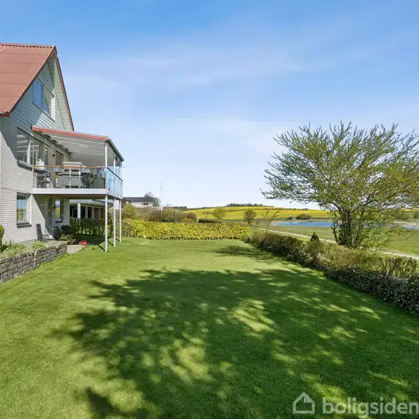 En hvid murstensbygning med rødt tag ligger ved siden af en velplejet græsplæne. En terrasse med stole ses på bygningen. Landskabet inkluderer marker og træer under en klar blå himmel.