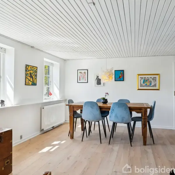 Spisebord med seks blå stole står i spisestue med hvide vægge og træloft. Rummet er oplyst af sollys fra tre vinduer, og kunstværker hænger på væggene.