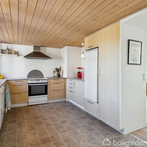 Et lyst køkken med træloft indeholder en ovn med emhætte, køleskab og køkkenborde. Dekorative elementer som frugter og køkkenredskaber supplerer det enkle design i rummet ved siden af.