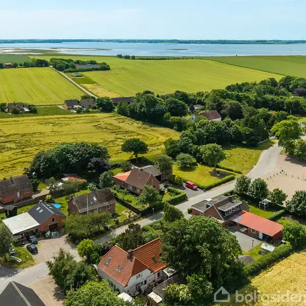 Huse står stille omgivet af grønne marker og træer, med en fjord i horisonten. Sollys belyser det landlige landskab.