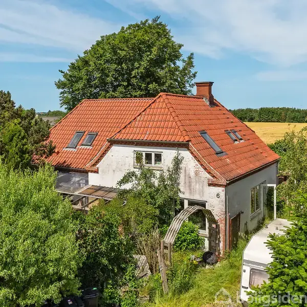 Et hvidt hus med rødt tegltag står blandt frodig vegetation. Sollys skinner på taget, og flere træer omgiver bygningen, med marker i baggrunden under en klar blå himmel.