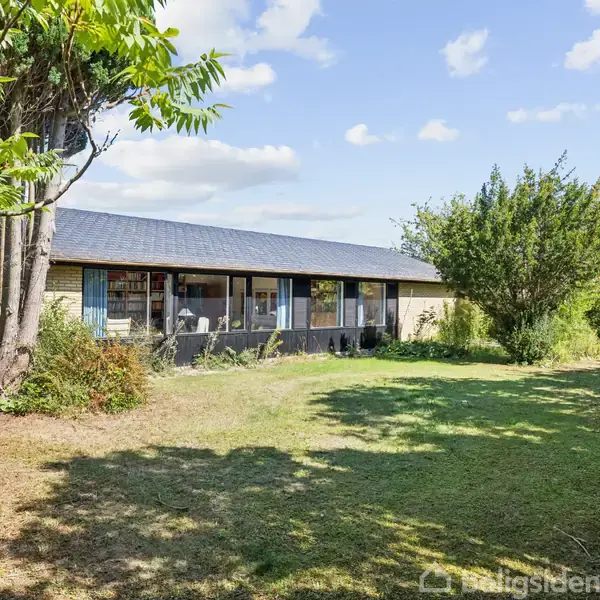 En etplans-bungalow med store vinduer står stille i en solrig have, omgivet af træer og buske, under en blå himmel med spredte skyformationer.