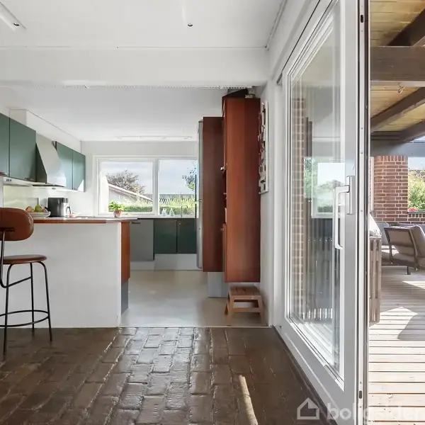 Køkken med grønne skabe og barstole ved en ø, åbner ud mod en terrasse med havemøbler. Store vinduer slipper lys ind og giver udsigt til haven.