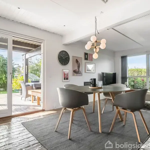 Rundt bord med fire stole står på gråt tæppe, der er placeret i et lyst rum med store vinduer. En moderne lysekrone hænger over bordet. Terrasse ses udenfor.