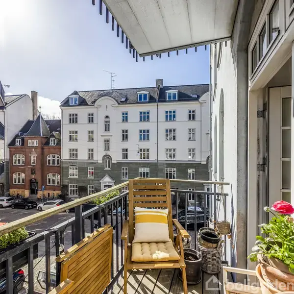 Balkon med træstol og planter står stille i sollys. Omgivet af klassiske bygninger i bymiljø.
