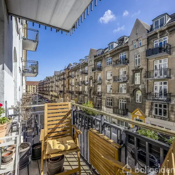 En balkon med to træstole og blomster i potter, overfor en række ældre lejlighedsbygninger under en klar blå himmel.