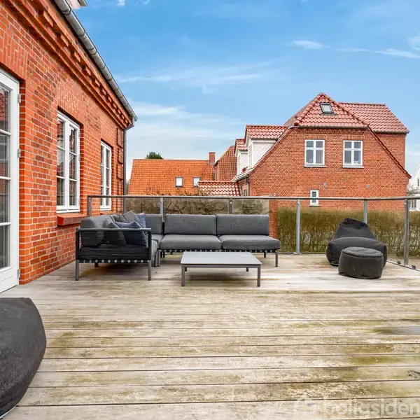 Terrassemøbler er placeret på en rummelig træterrasse med en rød murstensbygning i baggrunden. Sofaer og stole står ved et glasrækværk med udsigt over et villakvarter med flere bygninger.