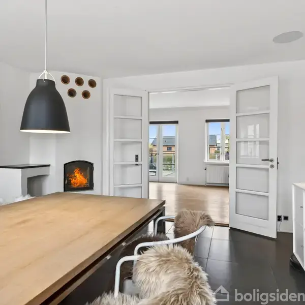 Spisebord med pelsbeklædte stole står i en rummelig, moderne stue. En pejs er tændt i baggrunden, omgivet af hvide vægge og dobbelt glasdøre, der fører til andet rum.
