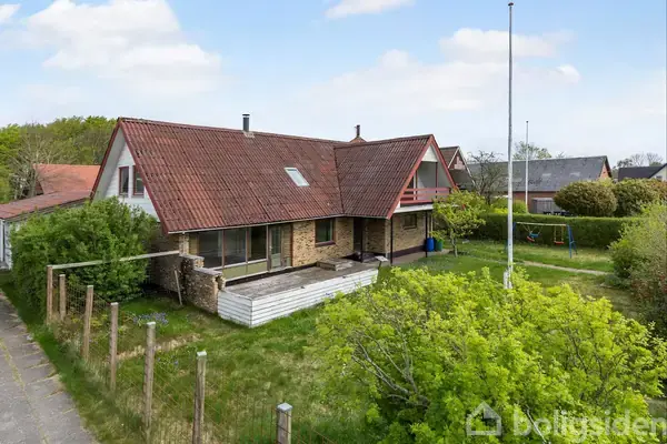 Et rødt hustag sidder på et hus med store vinduer og terrasse. Omgivet af grøn have og buske, er der en flagstang ved siden af. Tætliggende huse ses i baggrunden.