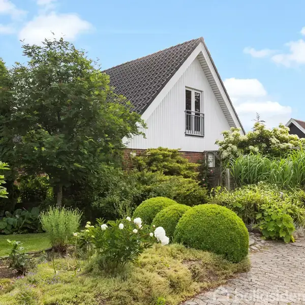 Et hvidt hus med en spids tagkonstruktion står omgivet af frodige grønne planter og buske. Foran huset er der en velplejet have. Himlen er blå med få skyer.