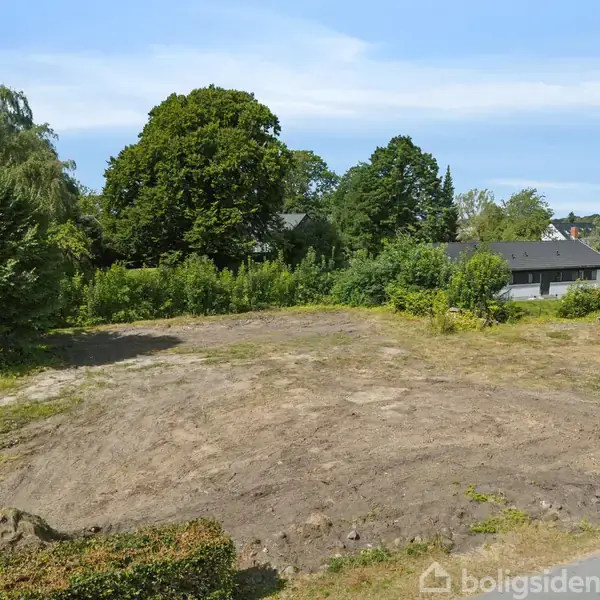 Et stykke bar jord ligger stille, omgivet af grønne buske og træer, med huse i baggrunden under en blå himmel.