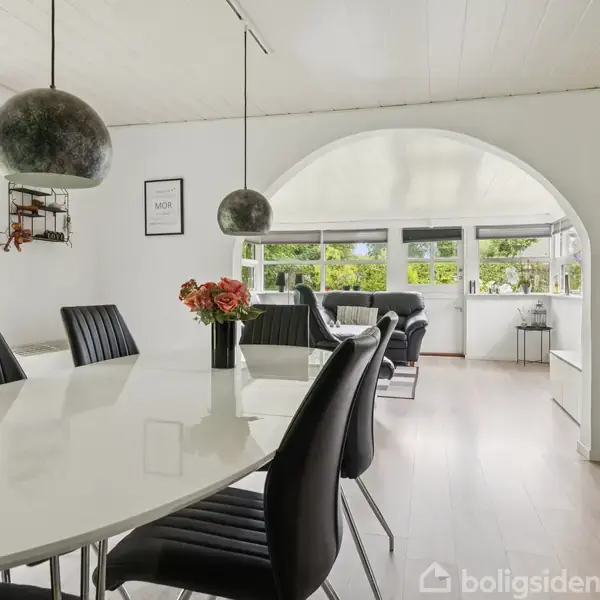Et ovalt, hvidt spisebord med sorte stole står i et lyst rum. Blomster pynter bordet. Store lamper hænger over. Der er en bue til en stue med store vinduer.