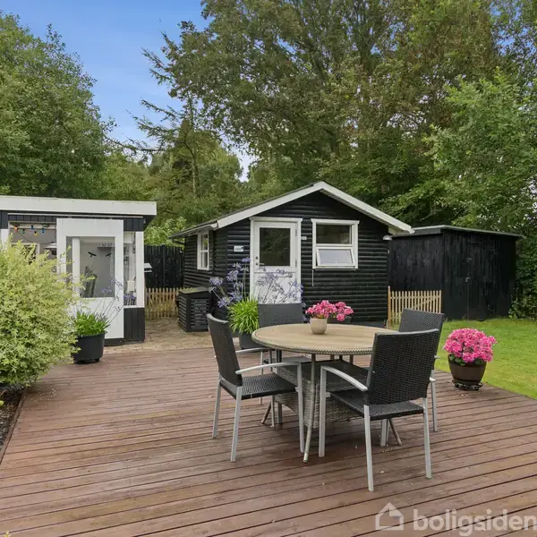 Et rundt havebord med fem stole står på en træterrasse. I baggrunden ses to små, sorte huse omgivet af grønne buske og græsplæne.