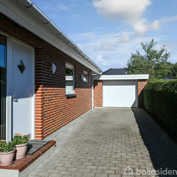 En murstensbygning med en hvid dør flankeret af potteplanter leder til en flisebelagt indkørsel mod en garage i baggrunden, omgivet af grønne hække og træer under en blå himmel.