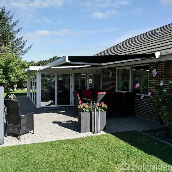 Terrasse med flisebelægning og udtrækkelig markise, omkranset af en murstensvæg. To stole og blomsterpotter står i solen. Haven har grønne græsplæner og buske.