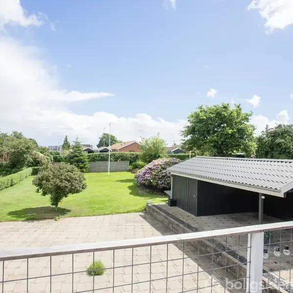 En have med en græsplæne er omgivet af forskellige træer og buske. En terrasse forrest rummer borde og stole. Til højre står en sort træbygning med et gråt tag.