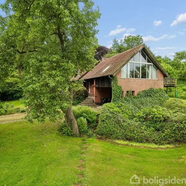 Hus med skråtag og store vinduer omkranset af frodig grøn vegetation. Trapper fører op til indgangen, og haven indeholder træer og buske under en blå himmel.