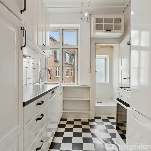 Køkkenet har hvide skabe og sorte greb, en sort bordplade, og flisebelagt væg med skakternet gulv. Et stort vindue giver udsyn til murstensbygninger udenfor.