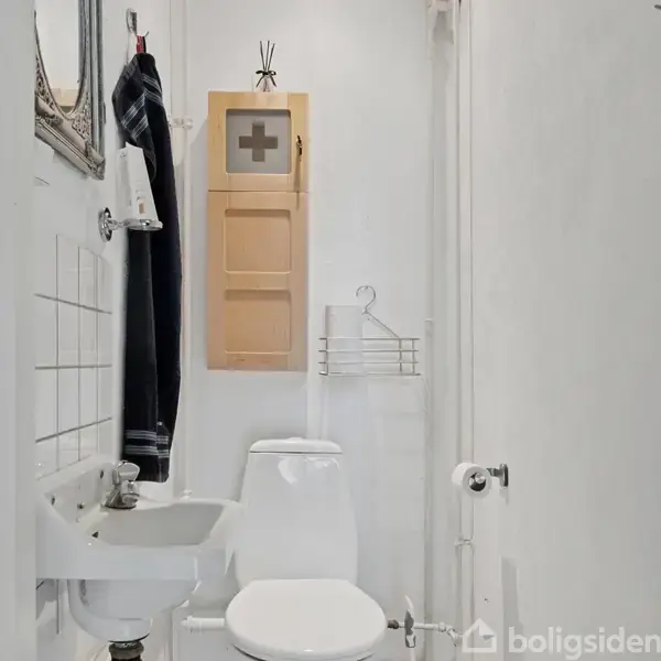 Et lille badeværelse har en hvid håndvask, et toilet og et træskab med et kryds på døren. Sorte håndklæder hænger ved siden af. Væggen er hvid.