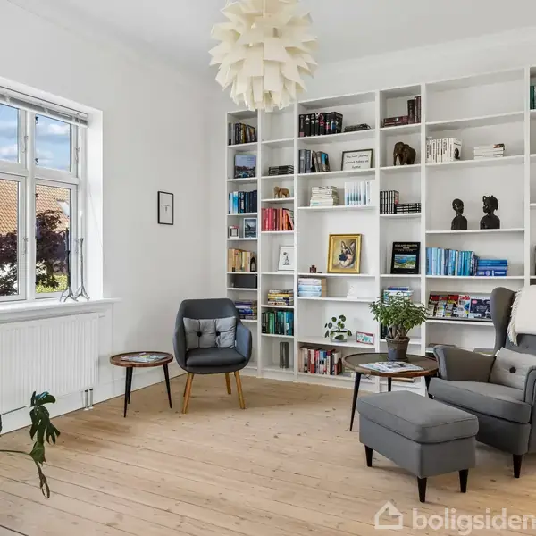 Reol fyldt med bøger og dekorative genstande, står mod en hvid væg. I nærheden sidder to stole med små borde i en lys og rummelig stue med planter ved vinduet.