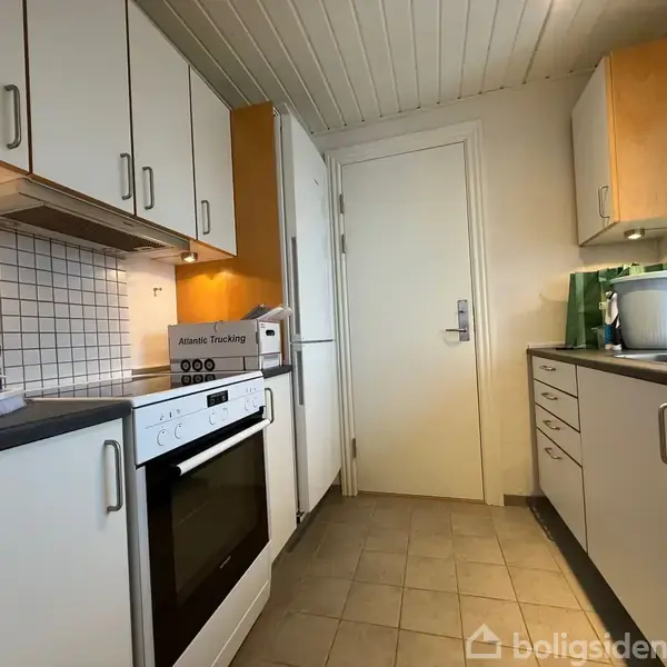 En køkken med hvide skabe og sorte bordplader har en ovn og en gryde på komfuret. Kontekst: Rummet har lys flisegulv og dør i baggrunden. Ingen tekst fundet.