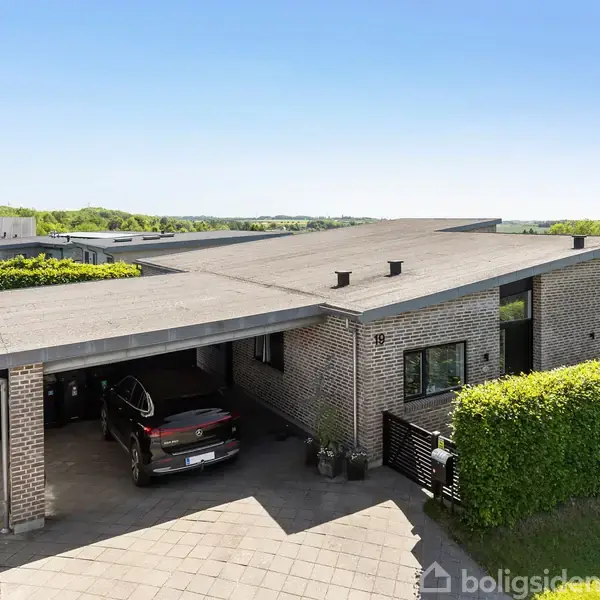 En moderne, lavbygget villa med fladt tag står stille. En sort bil holder i den tilhørende garage. Hække omkranser huset, der ligger i et grønt boligområde under en blå himmel.