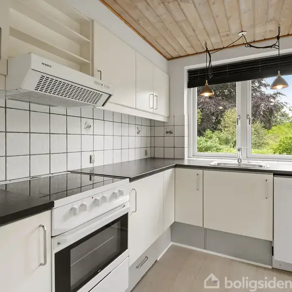 Komfur med emhætte er centralt placeret. Hvide skabe omkranser det i et moderne køkken. Ved vinduet er der en opvaskemaskine og vask, med udsigt til grønne træer.