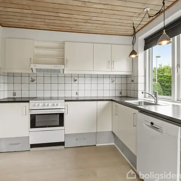 Køkkenets hvide skabe og opvaskemaskine er monteret langs væggen. En ovn står midt i, under en emhætte. Store vinduer til højre giver udsyn til grønne træer.