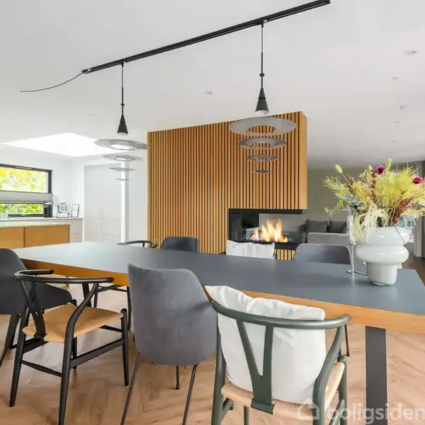 Et moderne spisebord står i et lyst rum med pejs og blomsterdekoration. Køkken i baggrunden med store vinduer, der giver rigeligt naturligt lys.