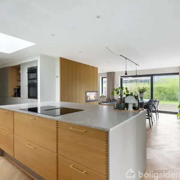 Køkken med træø, indbygget kogeplade og skuffer. Åben planløsning fører til spisestuen med glasdør mod haven. Moderne indretning med planteliv og kunst på væggen.