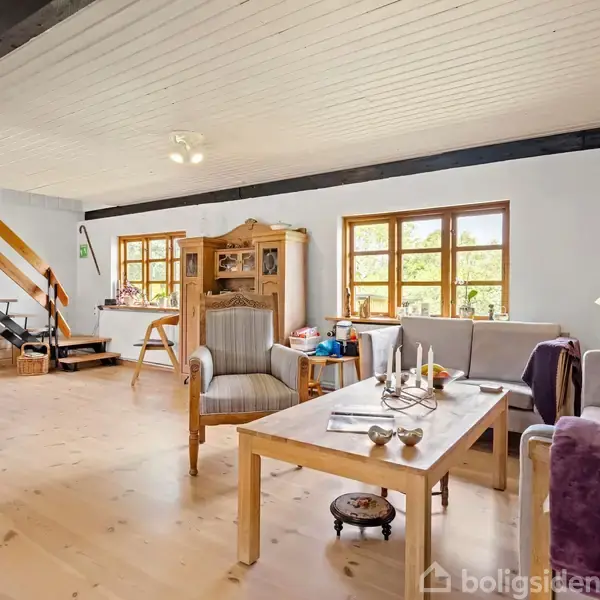 Stue med sofaarrangement og sofabord, hvor lys strømmer ind gennem vinduerne. Træloft og trægulv skaber en hyggelig atmosfære. En trappe leder til en øvre etage.